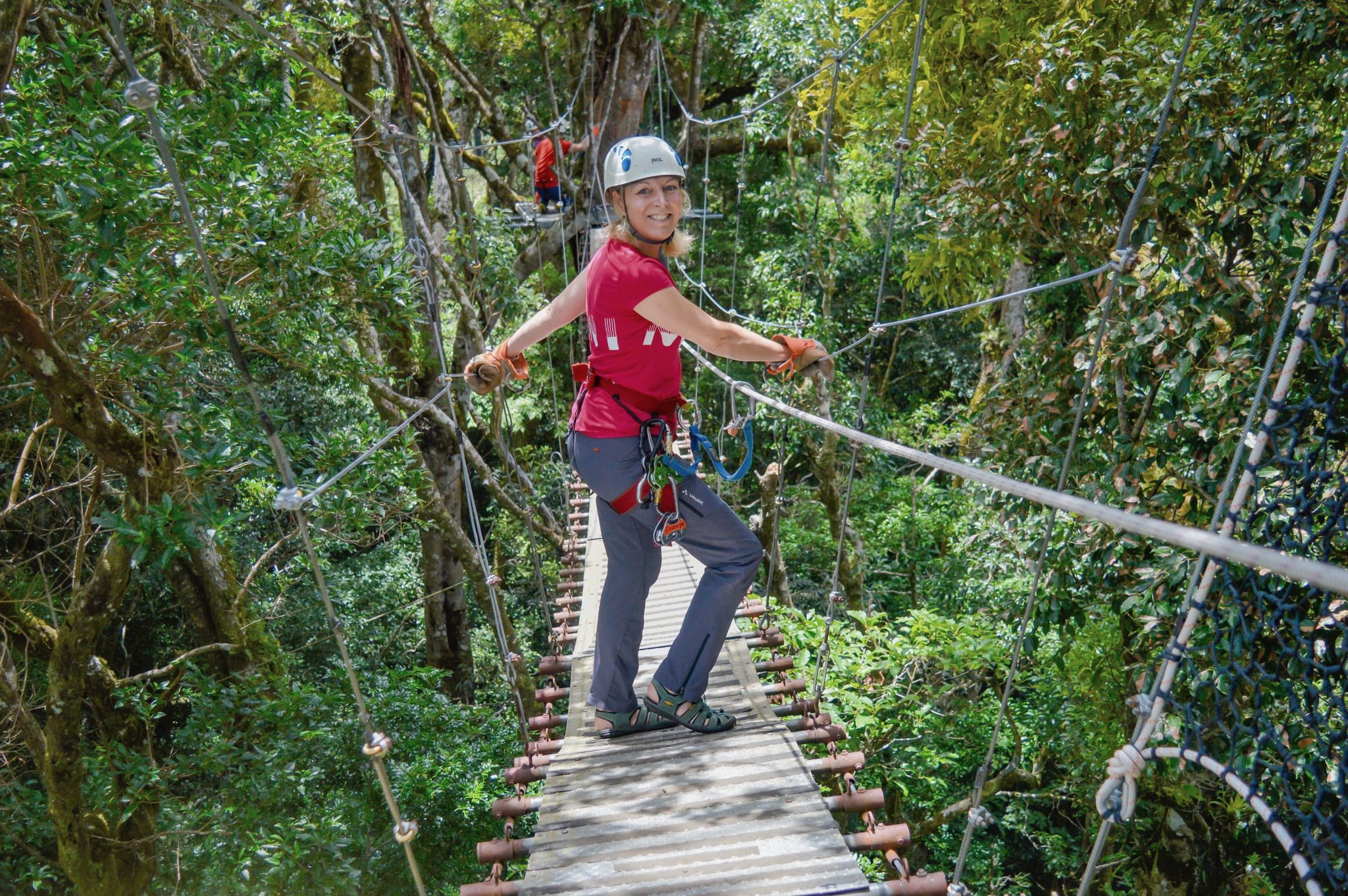 A visitor in a helmet, harness and gloves smiles back while crossing a suspension bridge in the cloud forest